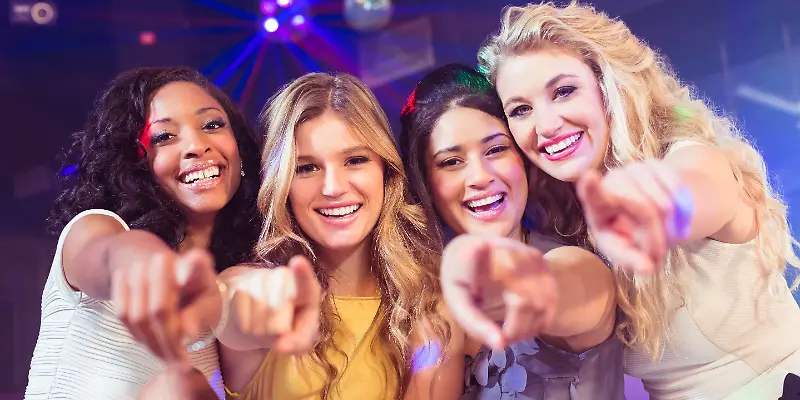 Vier lachende Frauen zeigen in die Kamera in einer Club-Atmosphäre.